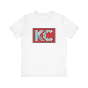 May include: White t-shirt featuring a red rectangle with the letters "KC" in teal. The letters have a double-lined design. The shirt is made of a soft material and has short sleeves.