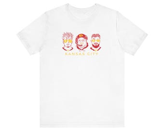 Op de afbeelding: Wit T-shirt met een rode en gele afbeelding van drie mannengezichten. De tekst "KANSAS CITY" is onder de afbeelding gedrukt.