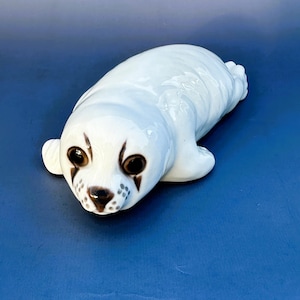 May include: A white ceramic figurine of a baby seal with brown eyes and a black nose. The seal is lying on its belly with its flippers outstretched.