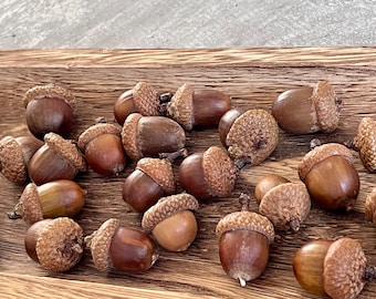 May include: A collection of brown acorns with textured caps, displayed in a wooden tray. The acorns vary in shades of brown, with some having a lighter hue. The tray has a natural wood grain.