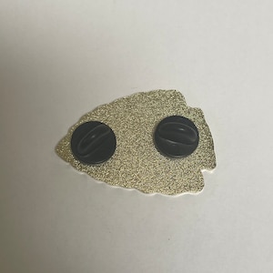 May include: Gold-colored metal pin in the shape of an arrowhead with two black rubber backings.