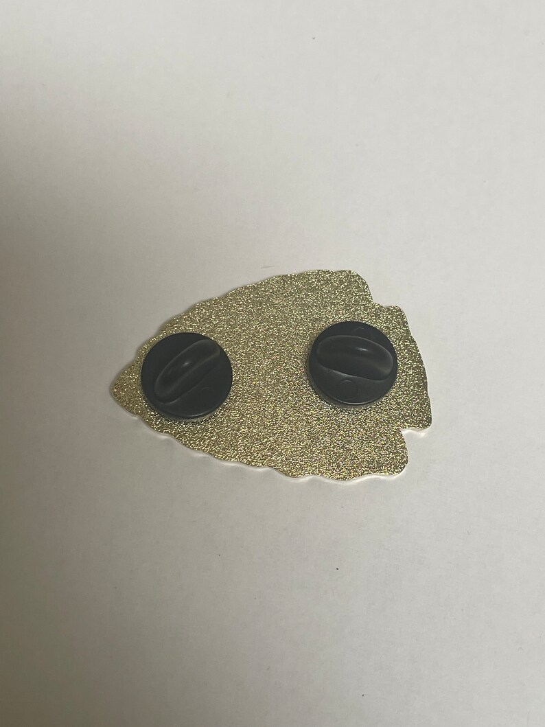 May include: Gold-colored metal pin in the shape of an arrowhead with two black rubber backings.
