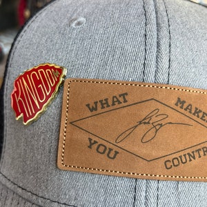 May include: A gray and black baseball cap with a red and gold pin that says "Kingdom" and a brown leather patch that says "What Makes You Country".