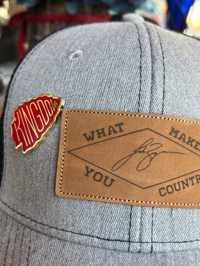 May include: A gray and black baseball cap with a red and gold pin that says "Kingdom" and a brown leather patch that says "What Makes You Country".