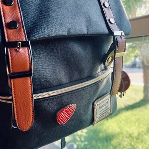 May include: A gray canvas backpack with brown leather straps and buckles. The backpack has a zippered pocket and a red enamel pin with the word "KINGDOM" on it. The backpack also has a small leather patch with the text "Brand Experience" and "WIZARDS" on it.