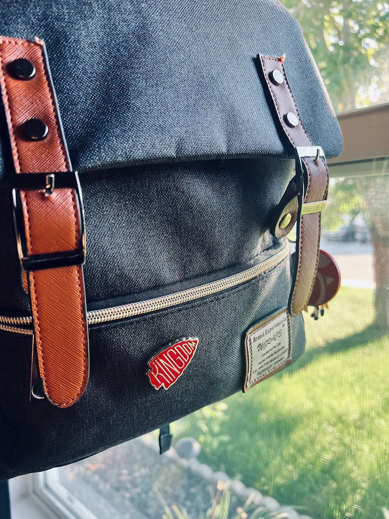 May include: A gray canvas backpack with brown leather straps and buckles. The backpack has a zippered pocket and a red enamel pin with the word "KINGDOM" on it. The backpack also has a small leather patch with the text "Brand Experience" and "WIZARDS" on it.
