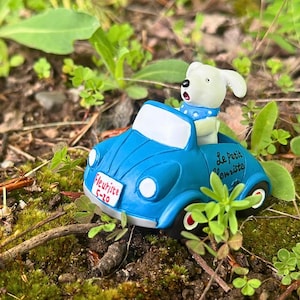 May include: A miniature blue car with a white windshield and a white dog figurine wearing a blue collar. The car has the text "Le Petit Fleuriste" and "5-20" on it. The car is surrounded by green plants and brown twigs.