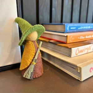 May include: A felt witch doll with a green hat, yellow cape, and a broom. The doll is next to a stack of books with titles including "The Cocktail" and "Hungry for More". The witch doll is a whimsical decorative item.