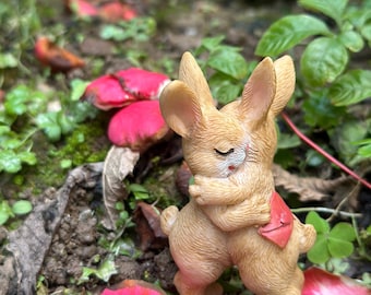 Może przedstawiać: Mała, brązowa, ceramiczna figurka królika z zamkniętymi oczami, która obejmuje siebie. Figurka stoi na łóżku z różowych płatków kwiatów.