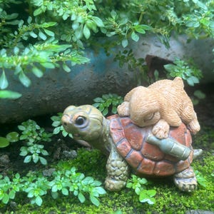 May include: A charming figurine of a tortoise with a sleeping rabbit on its back. The tortoise has a green head and legs, a brown shell, and a bottle strapped to its back. The rabbit is light brown. The figurine is surrounded by green plants.