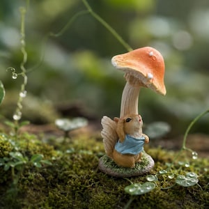 Op de afbeelding: Een klein beeldje van een lichtbruine eekhoorn in een lichtblauw hemdje, rechtopstaand onder een grote paddenstoel met een oranje hoed. De eekhoorn heeft een pluizige staart en kijkt omhoog naar de hoed van de paddenstoel met een gestructureerde steel.
