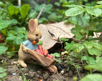 May include: A small, brown rabbit figurine wearing a blue shirt and a red bow tie pushes a wooden wheelbarrow filled with orange carrots. The figurine is set in a garden with green leaves and brown dirt.
