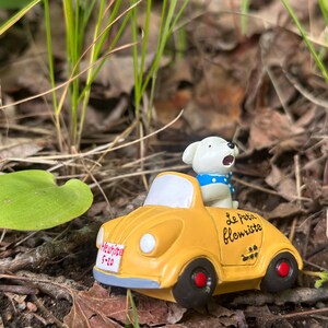 Puede incluir: Un coche de juguete amarillo con una figura de perro blanco que lleva un collar azul de lunares. El coche tiene ruedas negras con centros rojos y el texto "Le Petit Fleuriste" en el lateral. Una pequeña placa de matrícula dice "Heathipe 5-20".