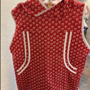 May include: A sleeveless red hooded vest featuring a repeating white heart pattern. The garment has white trim along the armholes, hem, and hood, with two vertical white-trimmed pockets on the front. A small tag with the number 12 is attached.