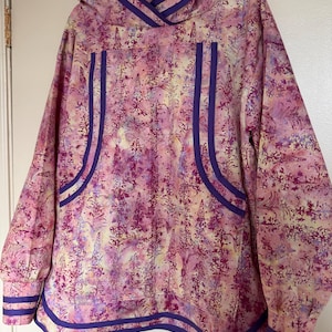May include: A long-sleeved hooded pullover featuring a vibrant pink, yellow, and purple floral pattern. The garment is accented with solid purple trim along the hood, the front pocket edges, and the hem, as well as double bands on the sleeve cuffs.