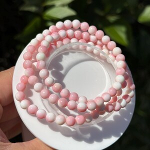 May include: A beaded bracelet featuring smooth, round beads in varying shades of soft pink and white. The beads are strung together in a circular pattern, displaying a gradient of light pink to white tones throughout the entire strand.