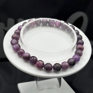 May include: A circular beaded bracelet featuring polished, spherical purple and dark violet gemstone beads. The beads are uniform in size and strung together in a continuous loop, displaying natural color variations and a smooth, reflective surface.