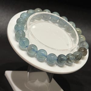 May include: A beaded bracelet featuring round, light blue aquamarine stones. The bracelet is displayed on a white, tiered stand against a dark background. The beads have a translucent quality, with subtle variations in color.
