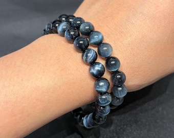 May include: Two bracelets made of polished, round, blue tiger eye stones. The bracelets are worn on a wrist, showcasing the natural variations in colour and pattern of the gemstones. The beads have a glossy finish.