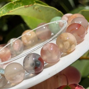 Puede incluir: Una pulsera de cuentas con piedras lisas, redondas y translúcidas en tonos melocotón, crema, rosa suave y verde apagado. Las cuentas presentan inclusiones y remolinos naturales, ensartadas en un cordón elástico transparente.