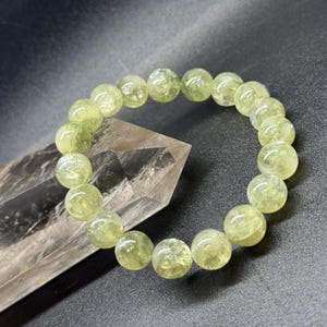May include: A light green beaded bracelet with round, translucent beads. The bracelet is resting on a clear, faceted crystal against a dark background. The beads have a subtle, natural variation in color.