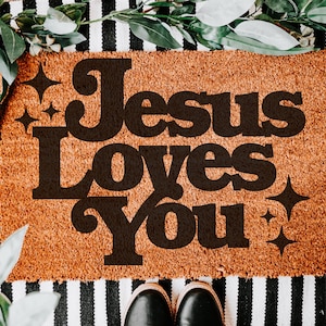Puede incluir: Una alfombra de coco marrón con el texto "Jesus Loves You" en letras estilizadas negras. El texto está rodeado de pequeñas estrellas blancas brillantes.