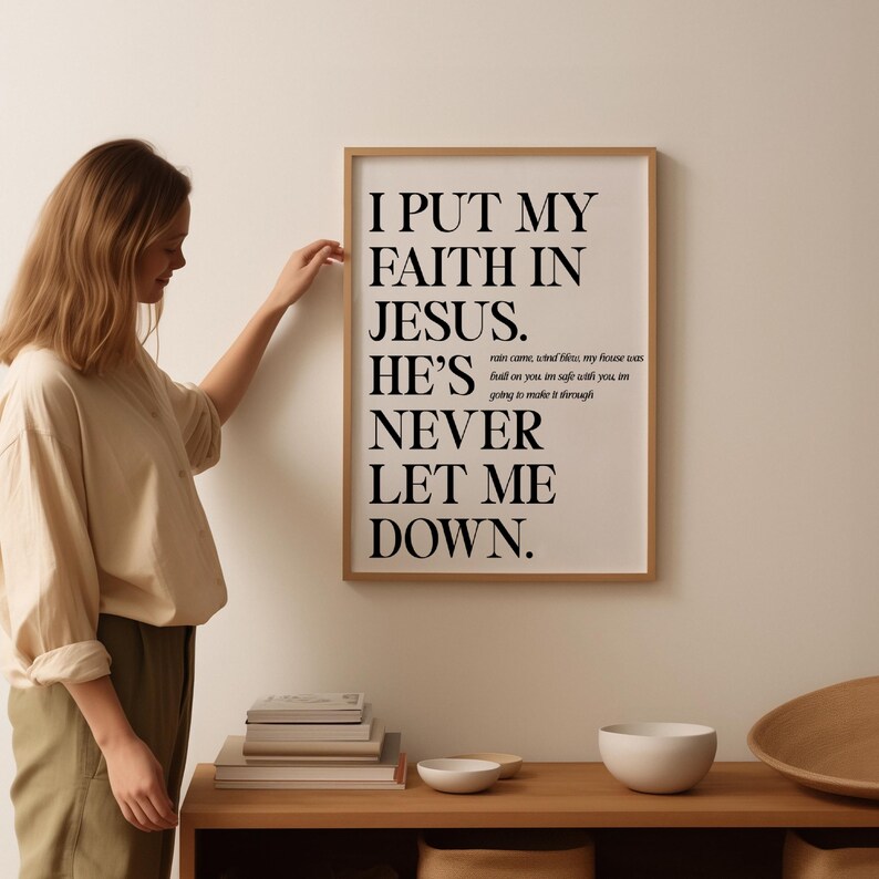 May include: A framed print with the text "I PUT MY FAITH IN JESUS. HE'S NEVER LET ME DOWN." in black font. The print is in a light brown frame and is hanging on a white wall. A person is touching the frame.