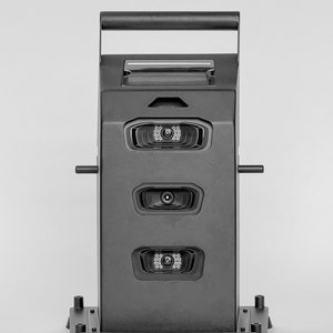 May include: A black, rectangular security camera system with three cameras mounted on the front. The system has a handle on the top and a base with four rubber feet.