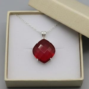 Può includere: Una collana d'argento con un grande pendente rosso scuro, sfaccettato e di forma quadrata. La collana è presentata in un portagioie beige aperto su sfondo grigio. Il pendente misura circa 2,5 cm di larghezza.