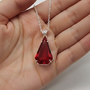 Op de afbeelding: Een zilveren ketting met een grote, facetgeslepen, driehoekige, dieprode hanger. De hanger is gezet in een zilveren montuur en hangt aan een fijne zilveren ketting. De ketting wordt gepresenteerd tegen een neutrale achtergrond.