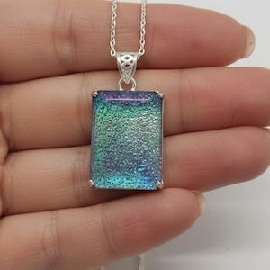 May include: A rectangular pendant necklace with a dichroic glass design. The pendant features a silver-colored frame and a chain. The glass has a textured surface with iridescent hues of blue, green, and purple.