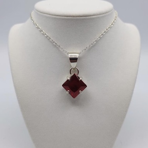 Può includere: Una collana d'argento con un pendente quadrato di pietra preziosa rosso scuro. Il pendente è incastonato in una montatura d'argento e pende da una delicata catena d'argento. La gemma ha una superficie sfaccettata, che riflette la luce. La collana è esposta su un manichino bianco.
