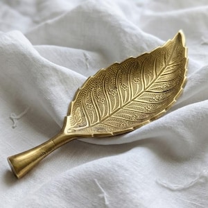 Op de afbeelding: Een gouden, bladvormige decoratieve schaal met ingewikkelde geëtste details. De schaal is gemaakt van metaal en rust op een witte, getextureerde stof. Het bladerdesign heeft gedetailleerde nerven en bloempatronen.
