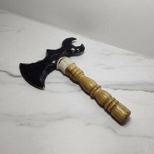 Op de afbeelding: Een zwarte en houten bijl met een gebogen blad en een houten handvat. De bijlkop is zwart en het handvat is lichtbruin. Het handvat is bij de bijlkop omwikkeld met een wit materiaal.