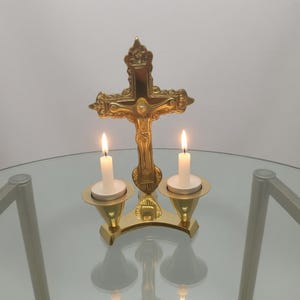 Op de afbeelding: Een goudkleurig kruisbeeld met een figuur van Jezus Christus, geflankeerd door twee brandende witte kaarsen in gouden houders. De kaarsen geven een warme gloed, het kruis is versierd.