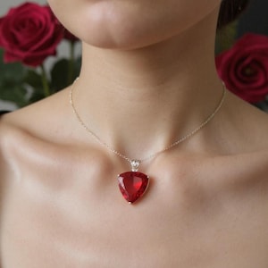 May include: A silver necklace featuring a large, triangular, ruby-red pendant. The pendant is set in a silver frame and hangs from a delicate silver chain. Red roses are visible in the background.
