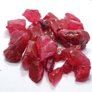 May include: A collection of rough ruby gemstones. The stones are a rich, deep red hue, with a slightly see-through quality. The shapes are uneven and angular, with visible facets and textures. The gemstones are scattered on a white surface.
