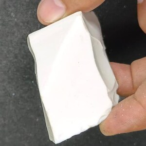 May include: A white, irregularly shaped piece of chalk is held between two fingers. The chalk has a rough, textured surface and appears to be freshly broken. The background is a dark, blurred surface.