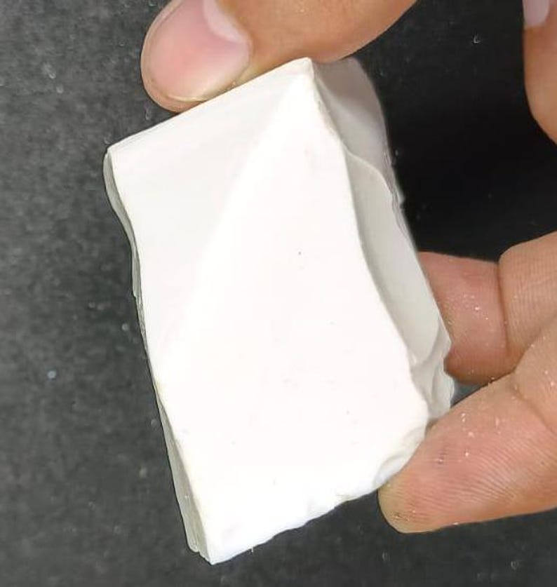 May include: A white, irregularly shaped piece of chalk is held between two fingers. The chalk has a rough, textured surface and appears to be freshly broken. The background is a dark, blurred surface.
