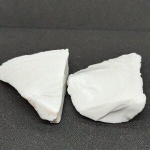 May include: Two irregularly shaped, opaque white rocks are displayed against a dark gray background. The rocks have a rough, textured surface and appear to be natural mineral specimens. The lighting highlights the subtle variations in the white color.