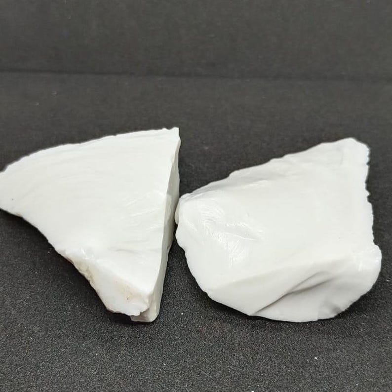 May include: Two irregularly shaped, opaque white rocks are displayed against a dark gray background. The rocks have a rough, textured surface and appear to be natural mineral specimens. The lighting highlights the subtle variations in the white color.
