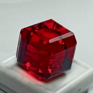 Può includere: Una pietra preziosa di colore rosso intenso tagliata a forma quadrata con bordi smussati. La pietra preziosa è su una superficie bianca.
