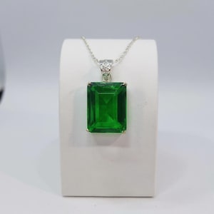 Puede incluir: Un collar de plata con un gran colgante rectangular de piedra preciosa verde esmeralda. El colgante está engastado en un marco de plata y cuelga de una delicada cadena de plata. La piedra preciosa tiene un corte facetado. El collar se exhibe sobre un soporte blanco.