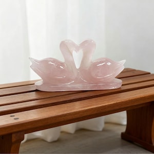 May include: A pink rose quartz sculpture of two swans facing each other, forming a heart shape with their necks. The swans are on a rectangular base. The sculpture is displayed on a brown wooden bench.