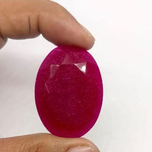 Peut inclure: Une pierre précieuse ovale et facettée d'un magenta vif. La gemme a une surface polie avec quelques facettes captant la lumière. La pierre est tenue entre deux doigts sur un fond blanc.