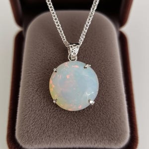 May include: A round, white opal pendant necklace with a silver chain. The opal has a faceted surface and is set in a silver bezel. The necklace is displayed in a brown velvet jewellery box.