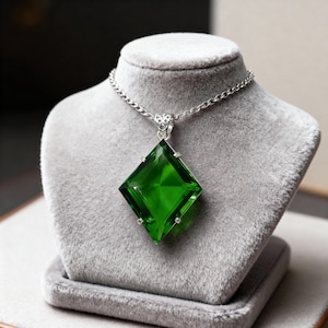 Puede incluir: Un collar con colgante de piedra preciosa verde vibrante. El colgante es una gran piedra verde esmeralda de corte cuadrado, engastada en un marco de metal plateado. La cadena del collar también es plateada y se exhibe sobre un busto de terciopelo gris.