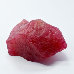 Peut inclure: Une pierre précieuse de rubis brute et non taillée, d'une teinte rouge profond. La surface de la pierre est texturée et irrégulière, avec un aspect légèrement translucide. Le rubis est posé sur un fond blanc uni.