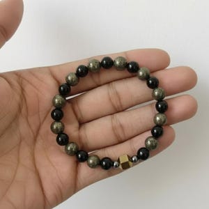 May include: A beaded bracelet featuring alternating black and gold-colored beads. The bracelet includes a gold-colored, geometric-shaped bead. The bracelet is shown on a hand.