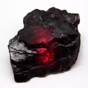 Puede incluir: Una gema áspera de color púrpura oscuro y rojo con un centro rojo intenso. La gema tiene una superficie texturizada y en capas.